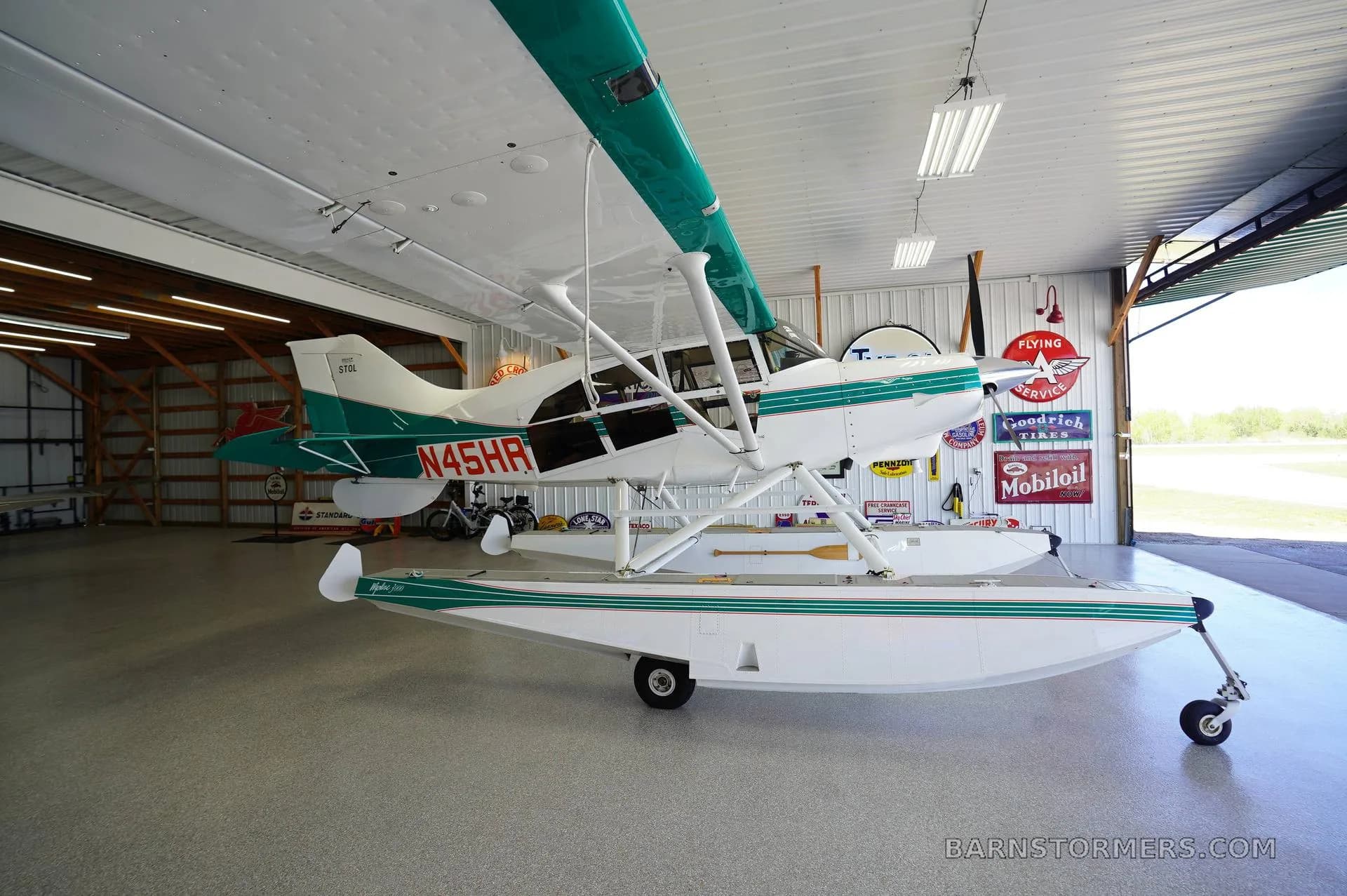 A white and teal floatplane, registration N45HR, is parked inside a hangar, viewed from the front-left.