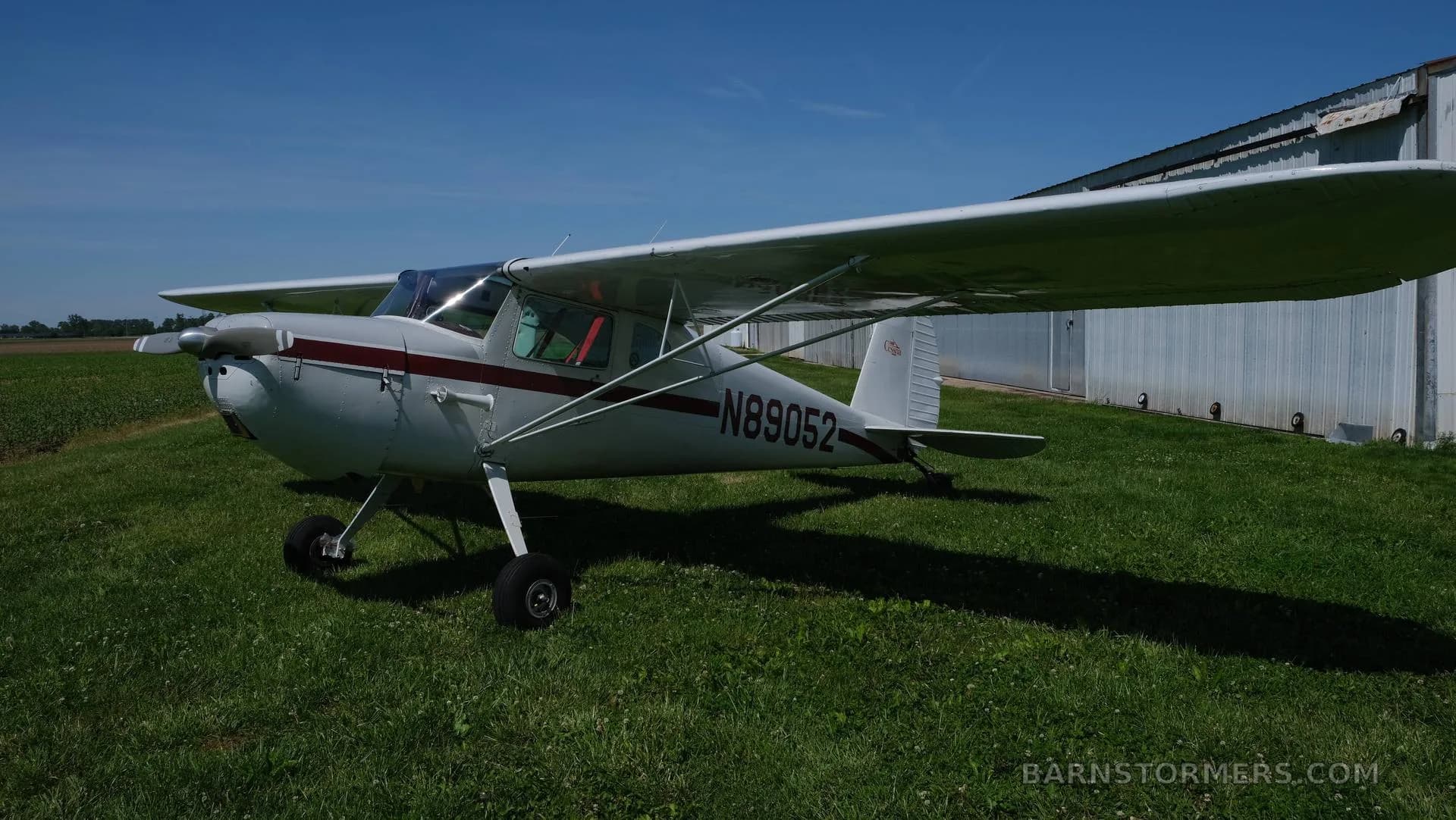 A front-quarter view of a white Cessna 170 with a red stripe, registration N89052, parked on green grass near hangars.