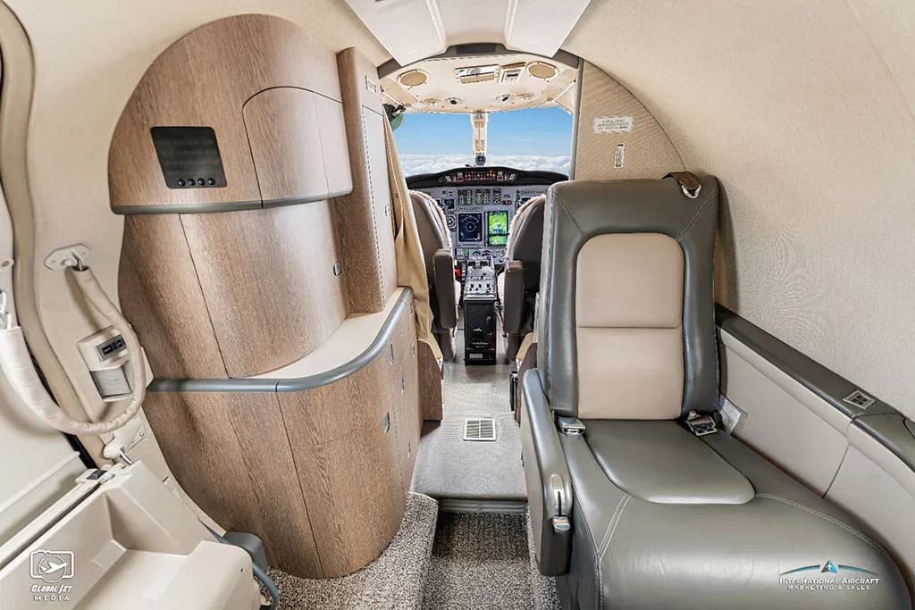A view from the business jet cabin looking forward, showing a wood-paneled galley area and the cockpit beyond.