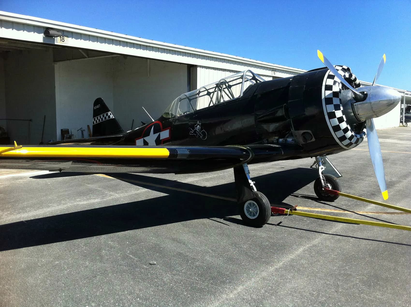 A black warbird aircraft with a checkered nose and yellow wing stripes, tied down on an airfield with a hangar in the background.