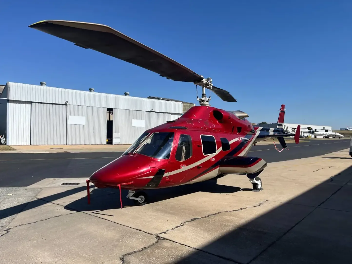 A red and white helicopter sits on the tarmac in front of a hangar.