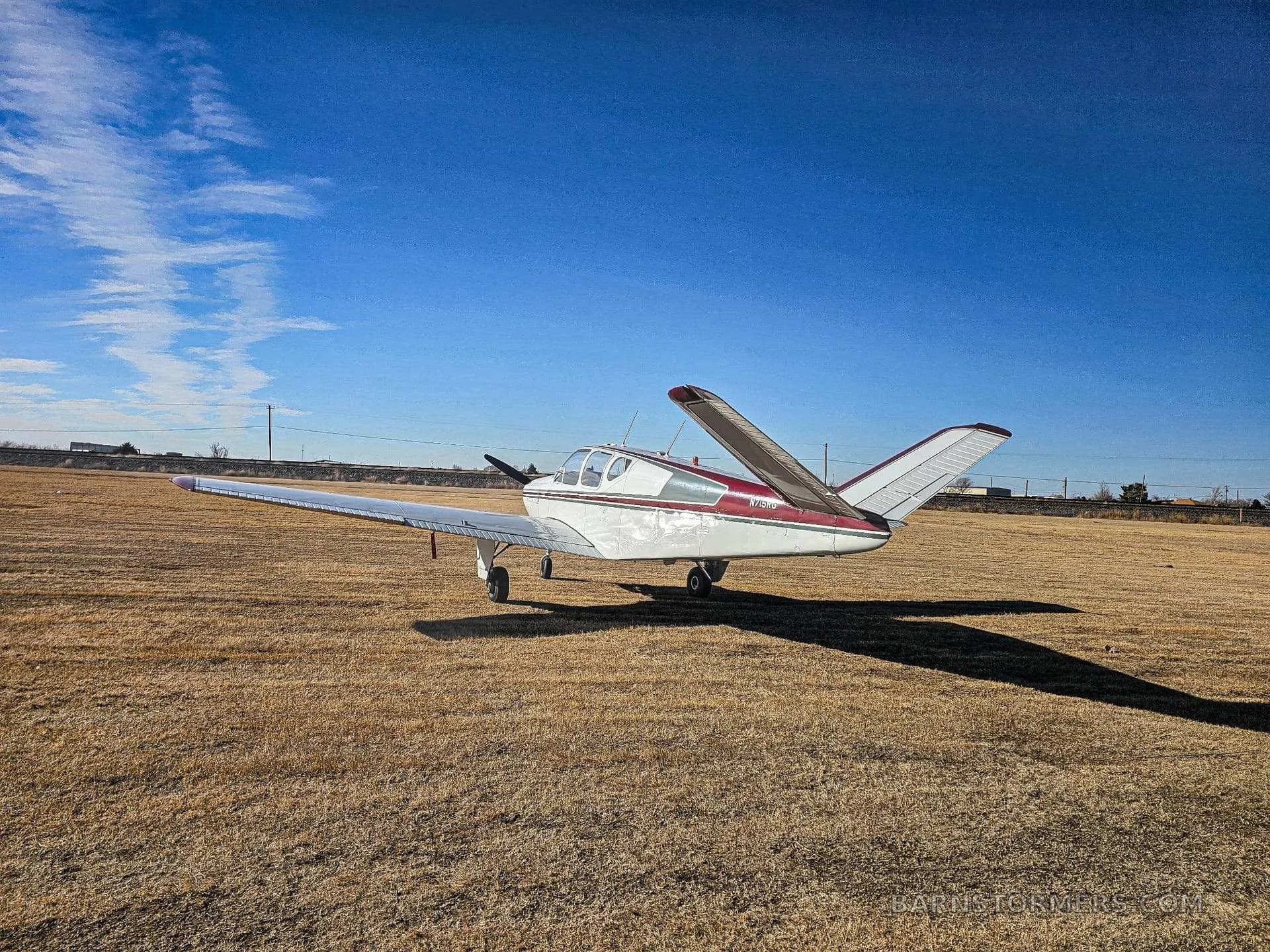 A white and red Mooney M20 series aircraft with its wings folded, parked on a grassy field under a blue sky.
