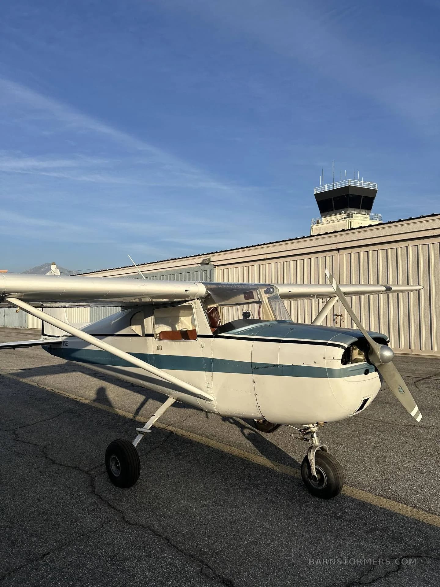 A white and blue Cessna 150/152 aircraft is parked on a tarmac with an air traffic control tower in the background.