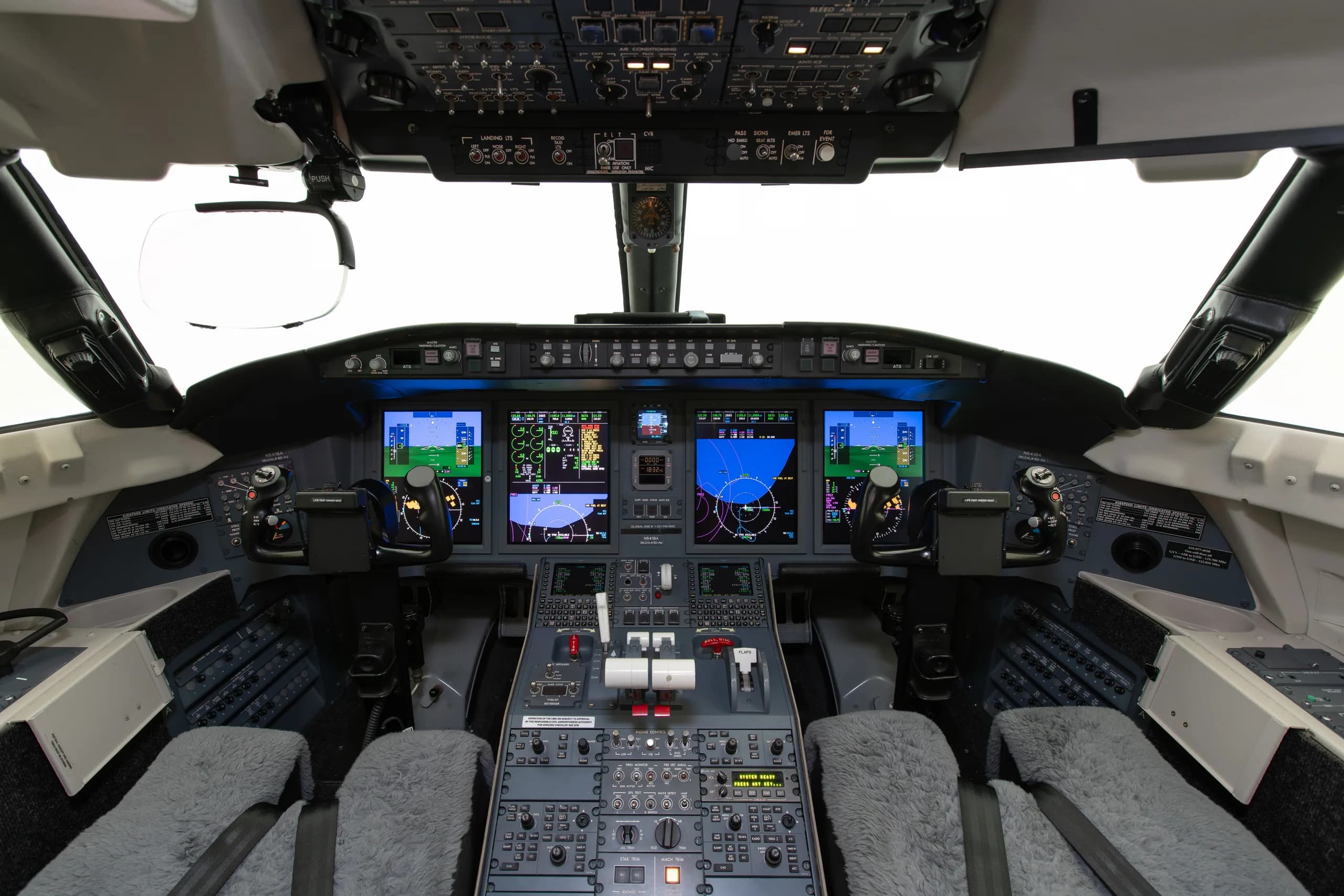 The cockpit of a modern business jet displays multiple digital screens, control panels, and two pilot seats with sheepskin covers.