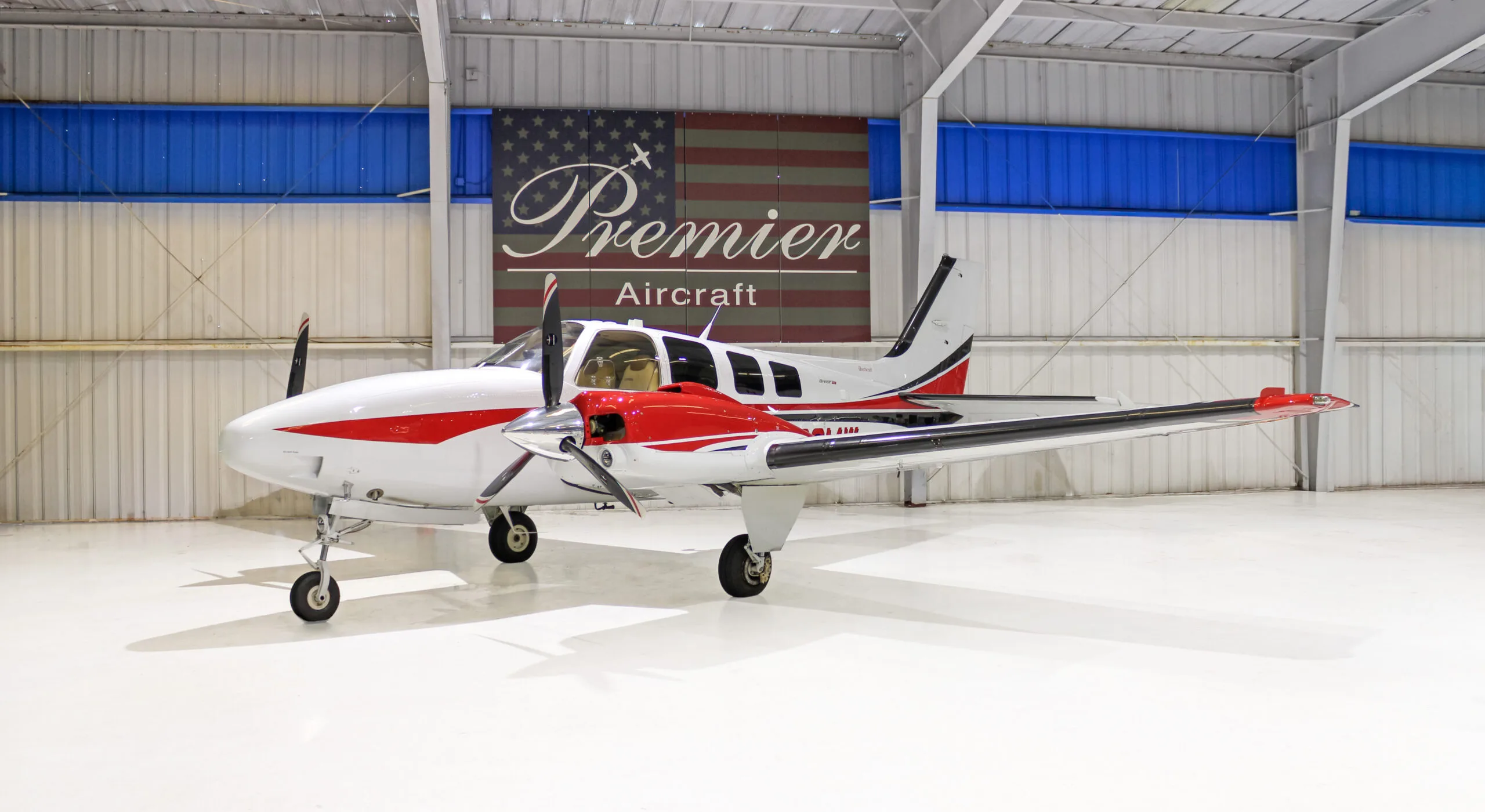 A white and red twin-engine propeller aircraft, a Beechcraft Baron, parked in a hangar.