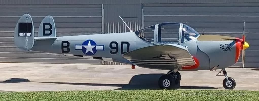 A grey single-engine aircraft with a red nose and "Sassy Baby" text is parked on a concrete surface next to grass.