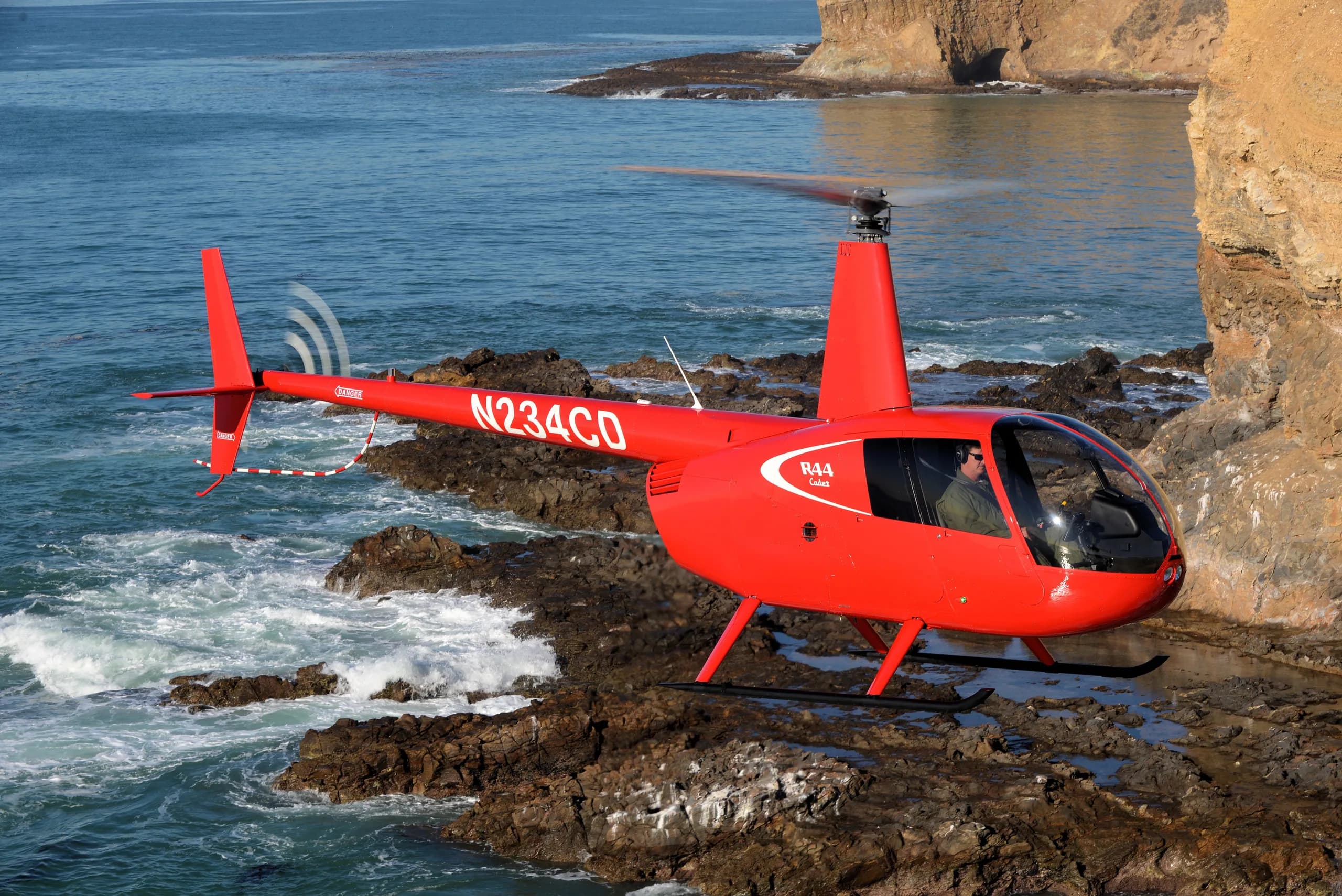 A red Robinson R44 helicopter flying near the ocean.