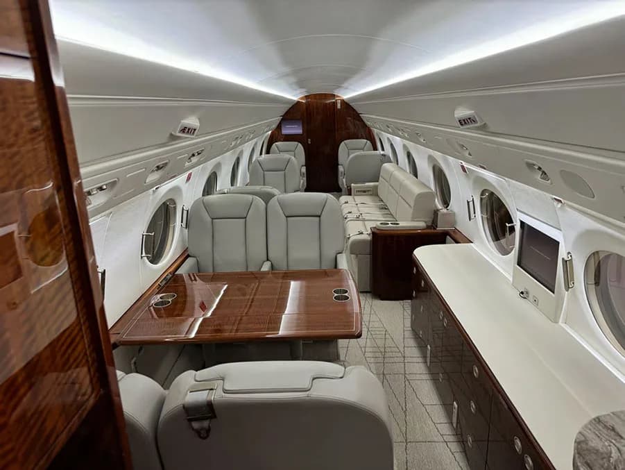 A luxurious aircraft cabin with light gray leather seats, a wooden table, and dark wood cabinetry.