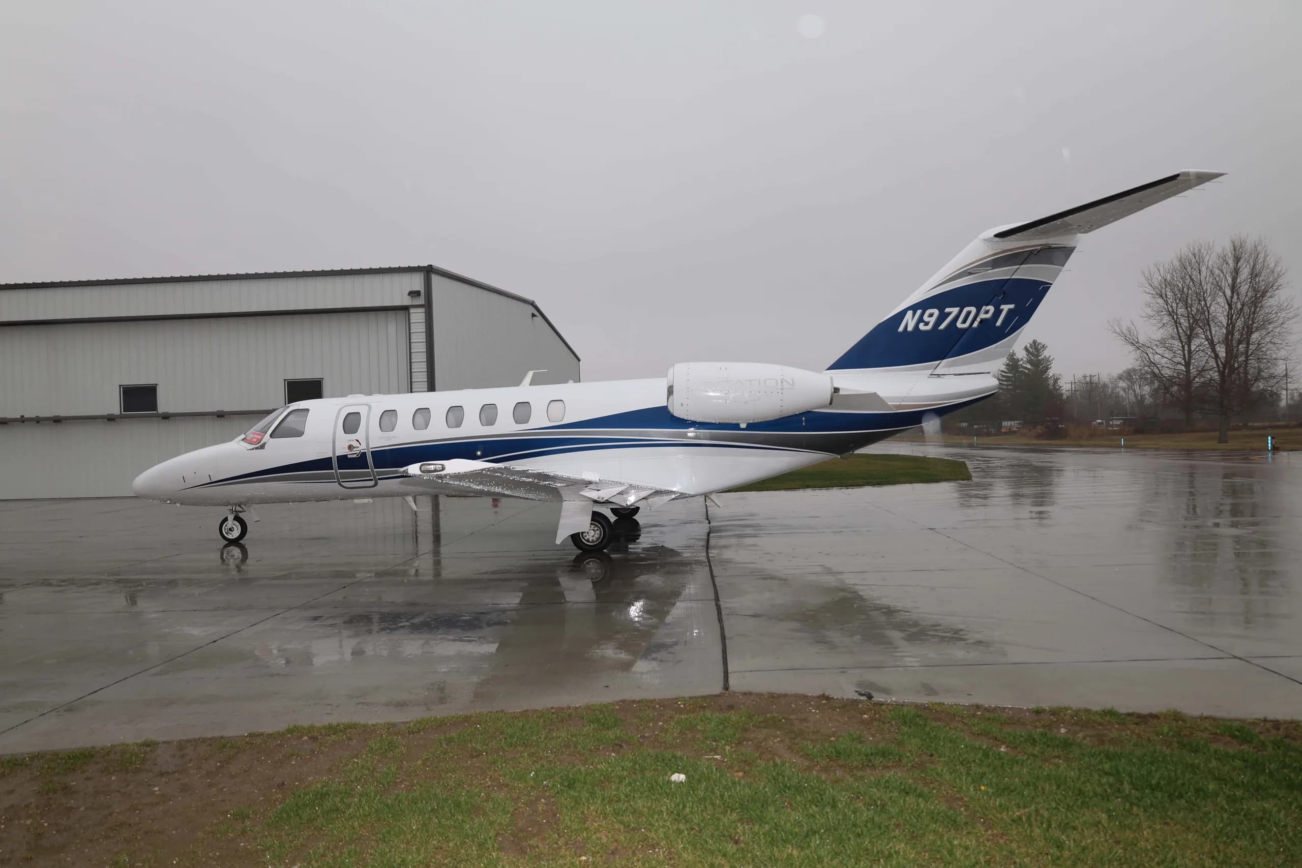 A white Cessna Citation M2 business jet with blue and grey stripes, registration N970PT, parked on a wet tarmac with a hangar in the background.