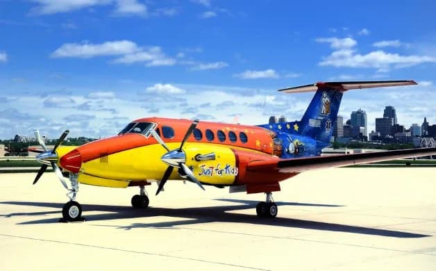 A colorful Beechcraft King Air aircraft with a 'Just for Kids' livery parked on a tarmac with a city skyline in the background.