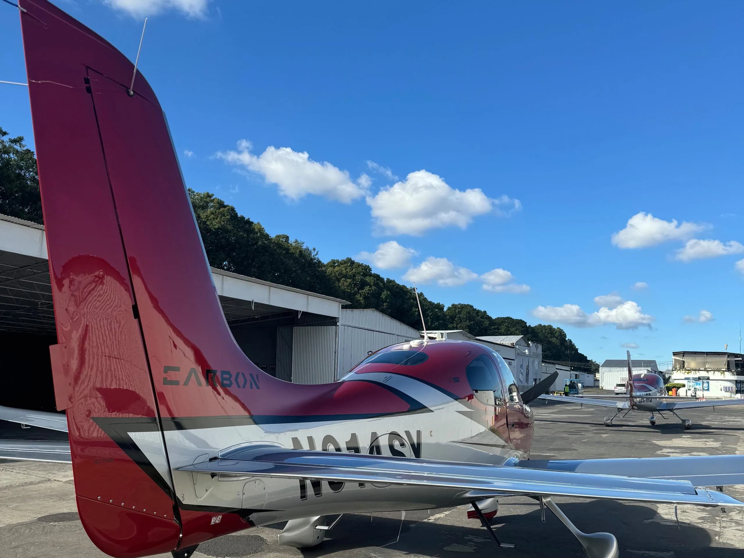 A rear view of the red and silver Cirrus SR22 aircraft N914SY parked on an airfield with other aircraft in the distance.