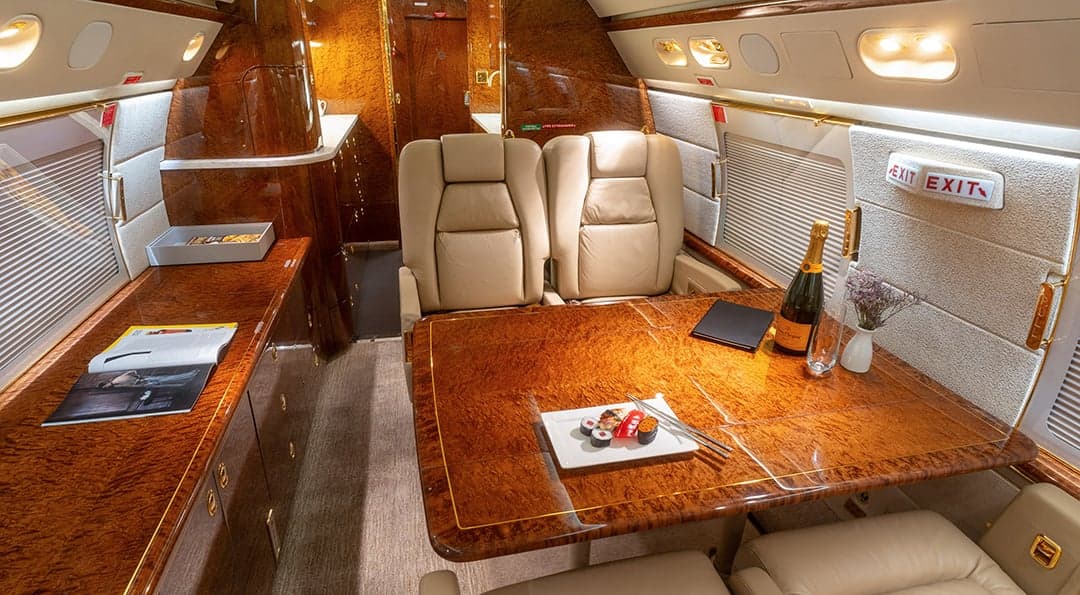 A private jet dining area with a polished wooden table set with sushi and champagne, surrounded by beige leather seats.
