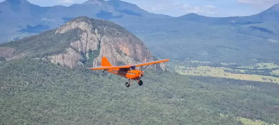 An orange high-wing aircraft flying over a lush green valley with large rocky mountains in the background.