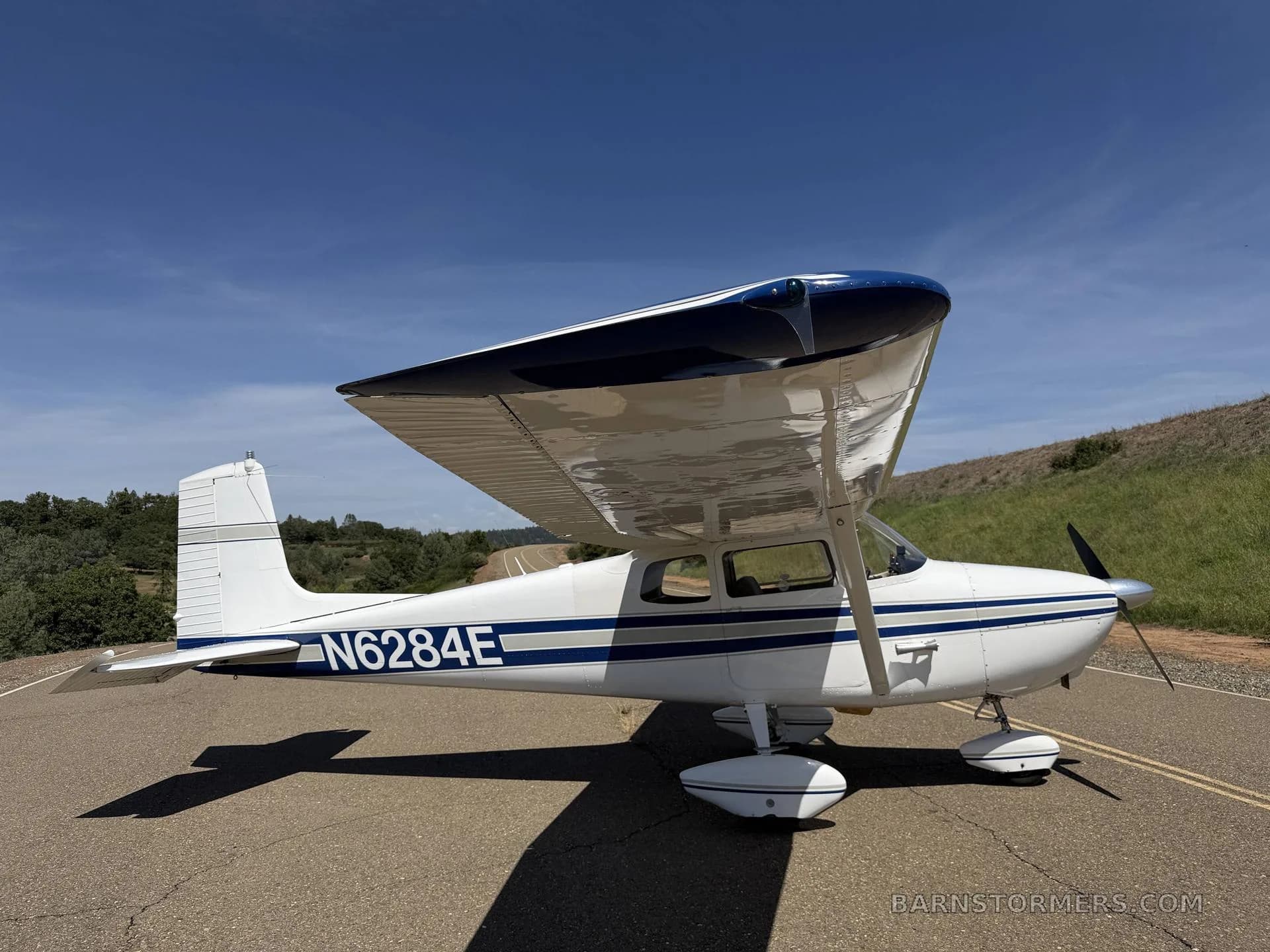 A white and blue single-engine high-wing aircraft, N6284E, parked on an asphalt strip under a clear blue sky, viewed from the front-right.