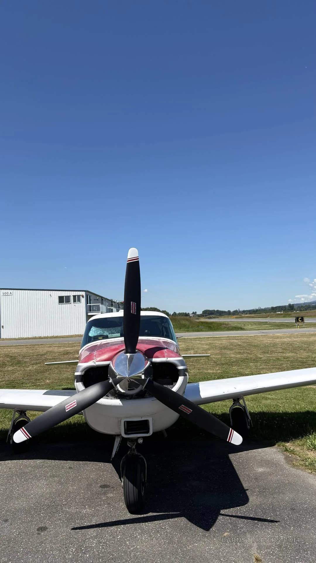 Front view of a white and red single-engine propeller aircraft with a three-blade propeller, parked on an airfield under a clear blue sky.