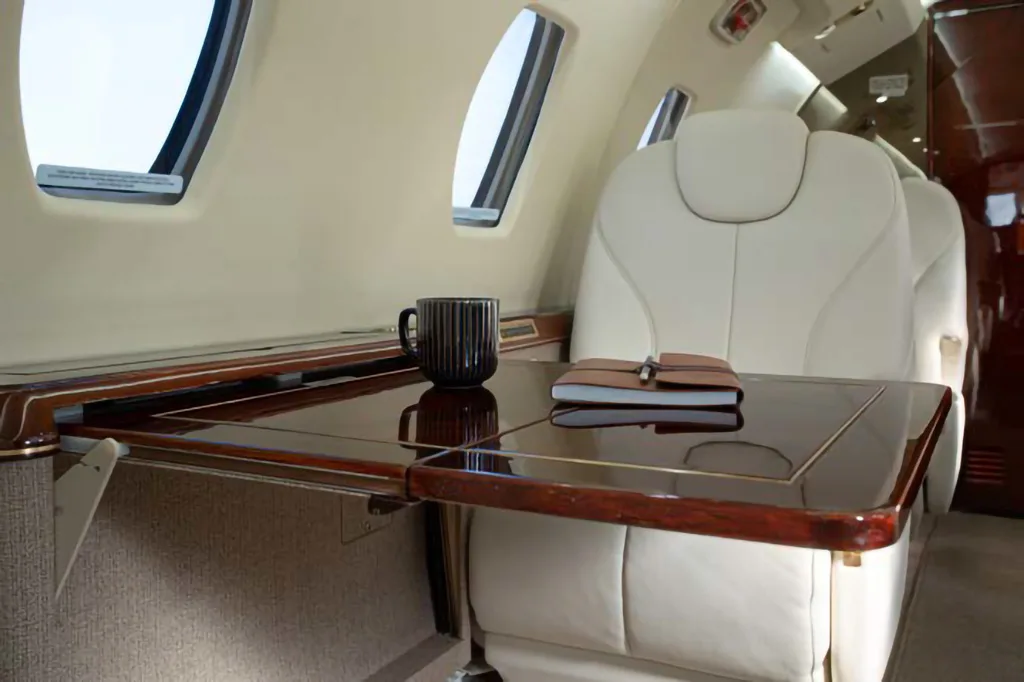 Close-up of a polished wooden table with a mug and notebook next to a cream leather seat in the aircraft cabin.
