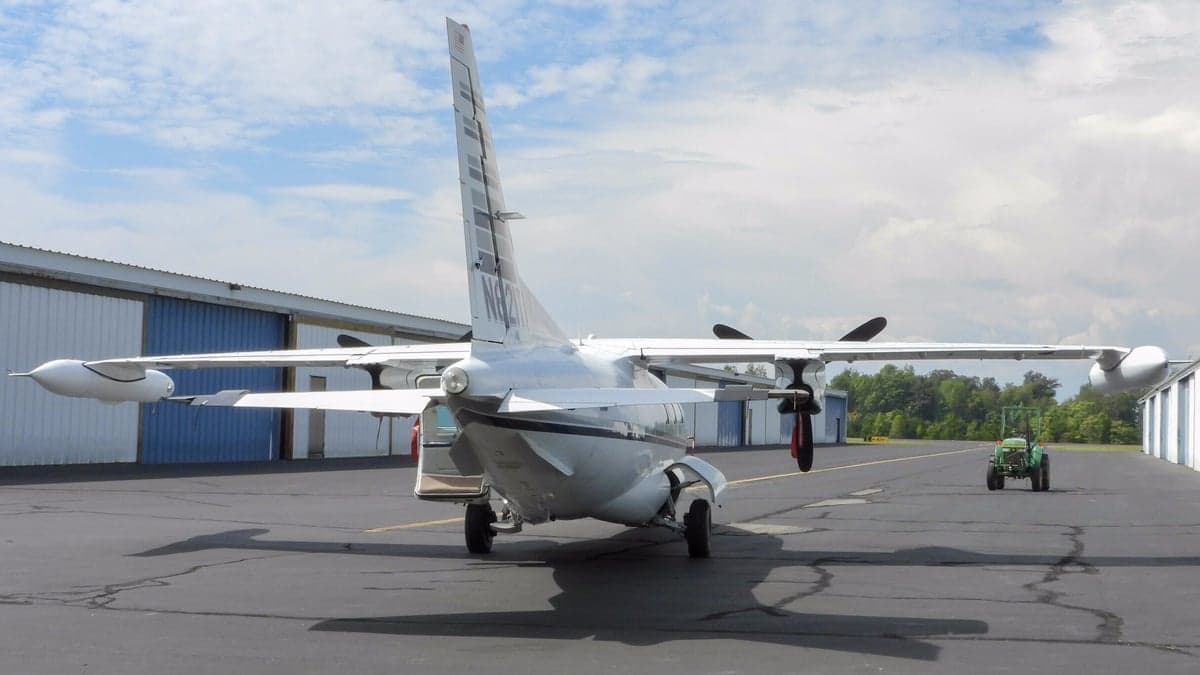 A white Mitsubishi MU-2 aircraft with its tail facing the viewer, parked on an airfield with hangars in the background.