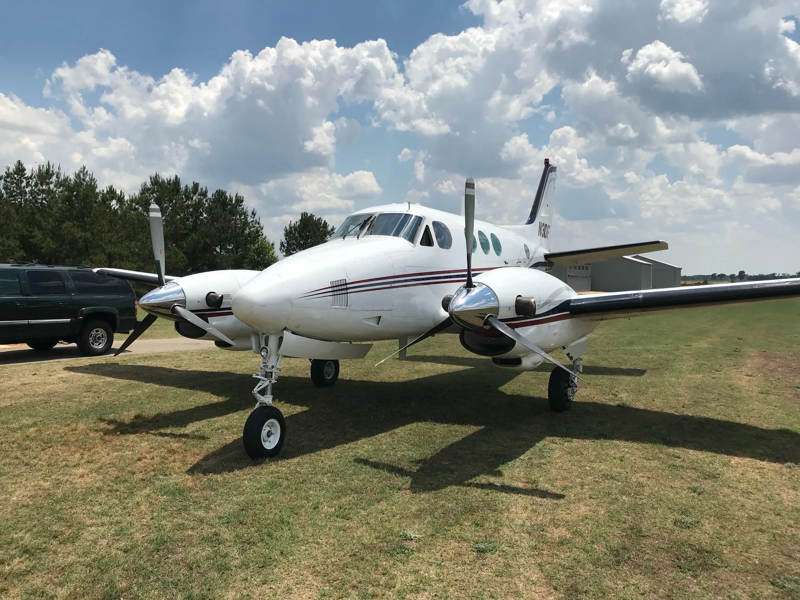 A white twin-engine turboprop aircraft with red and blue stripes is parked on a grassy field under a partly cloudy sky.