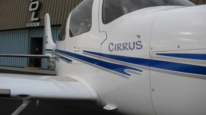 Side view of a white Cirrus aircraft with blue and grey stripes parked in front of a hangar.