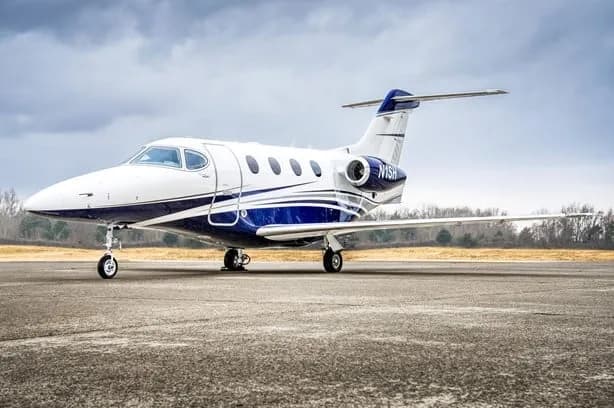 A white and blue business jet, tail number N1SH, parked on an airfield with trees in the background.