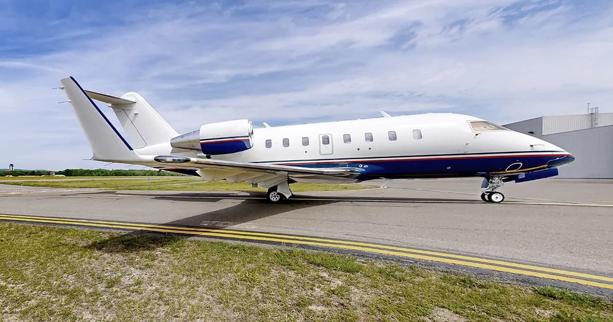 A white and blue Bombardier Challenger business jet is parked on a tarmac with a hangar in the background.