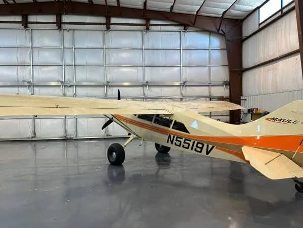 A Maule aircraft with registration N5519V, painted white and orange, viewed from the rear left in a hangar.