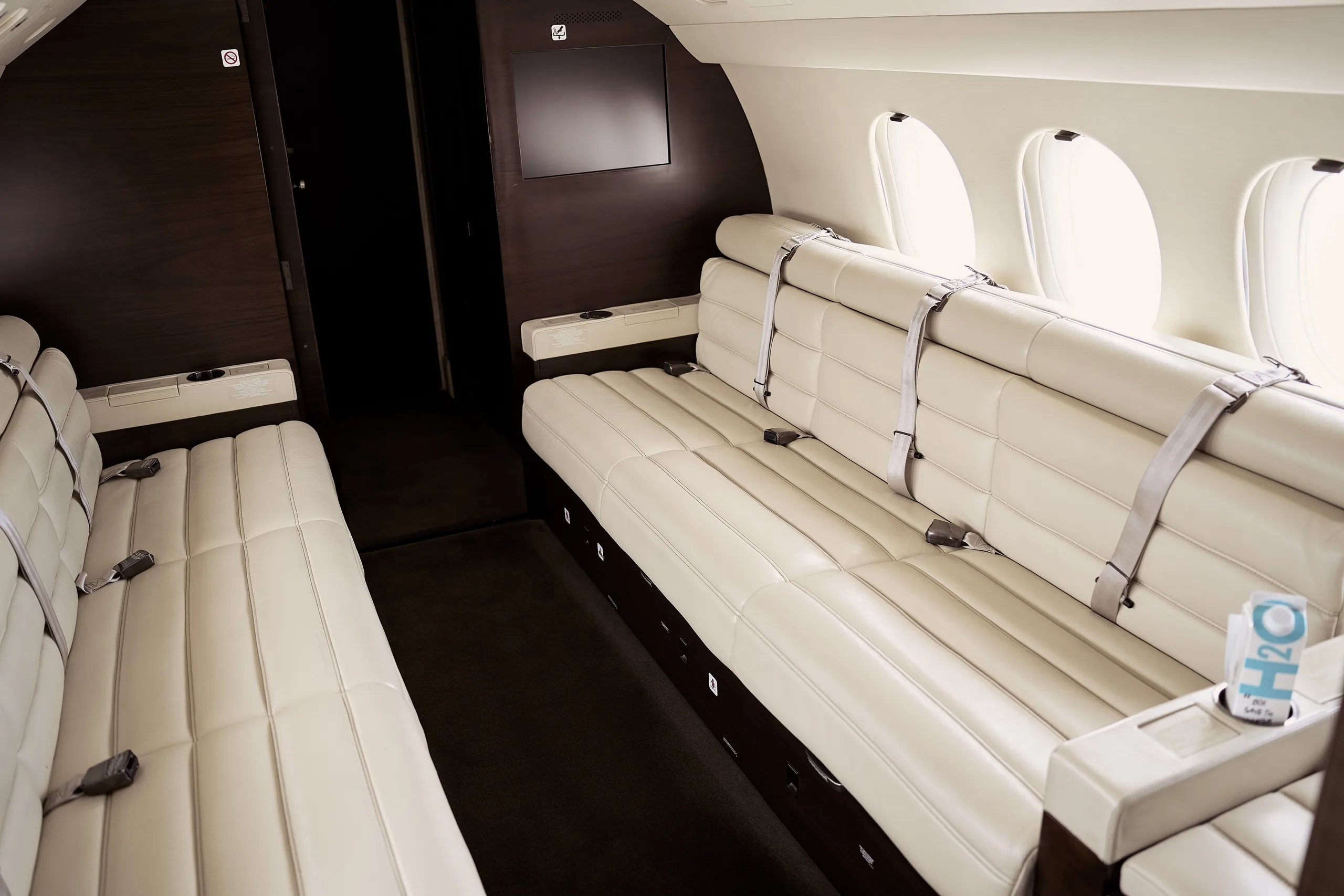A luxurious private jet cabin featuring long cream leather bench seats and a dark wood wall with a mounted screen.