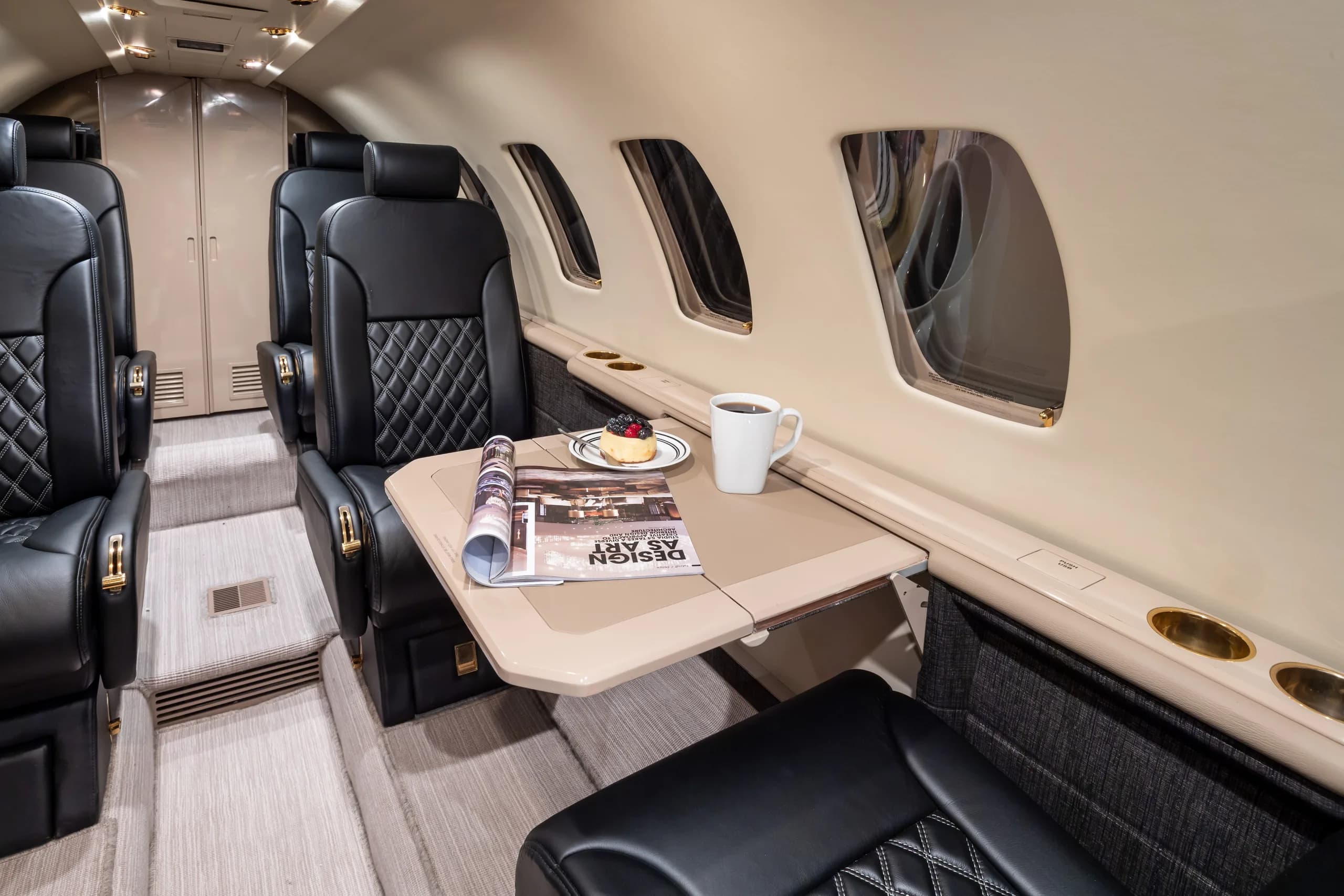 A side view of the business jet's cabin interior, highlighting a table with a magazine, coffee, and a pastry, next to black quilted leather seats.