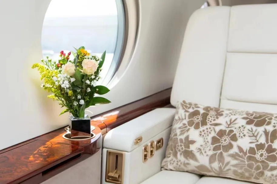 A close-up of a private jet interior showing a white leather seat, a window, and a vase of flowers on a wood-trimmed table.