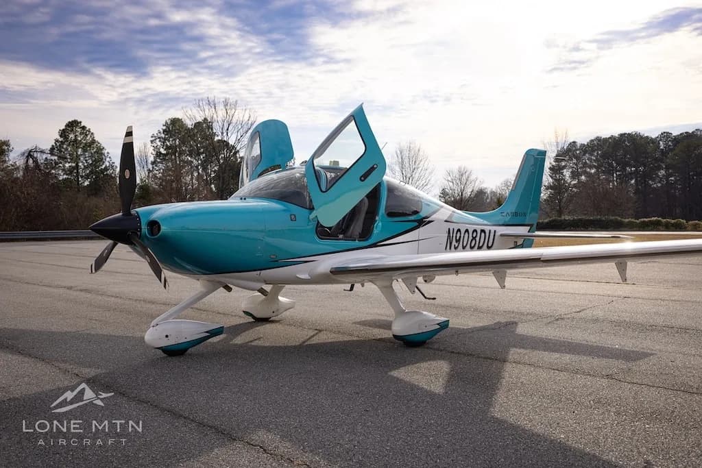 A teal and white Cirrus SR22 aircraft, N908DU, with its doors open, parked on an airfield.
