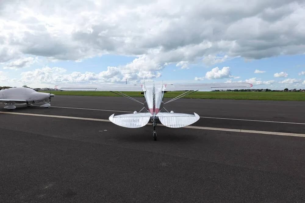 A direct rear view of a silver Cessna 170B with red wingtips, parked on an airfield next to another aircraft.