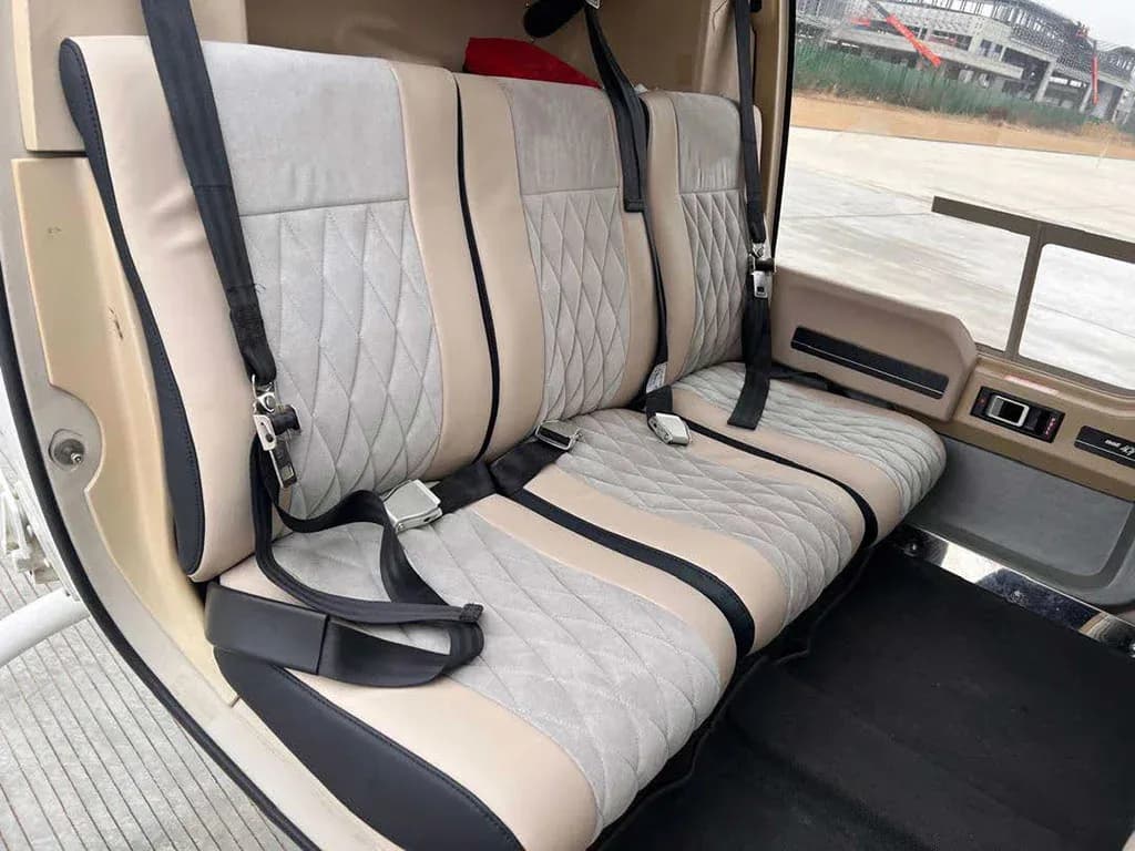 A side view of the helicopter cabin interior, showing a row of three beige and gray quilted seats with black seatbelts.