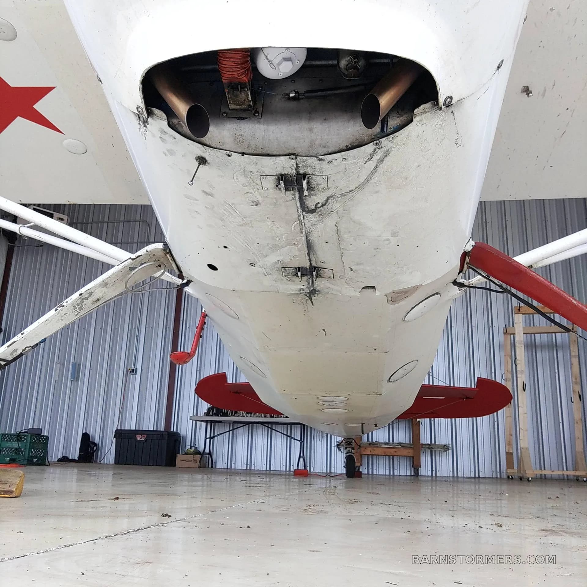 An underside view of a red and white Citabria aircraft, showing the exhaust pipes and landing gear, inside a hangar.
