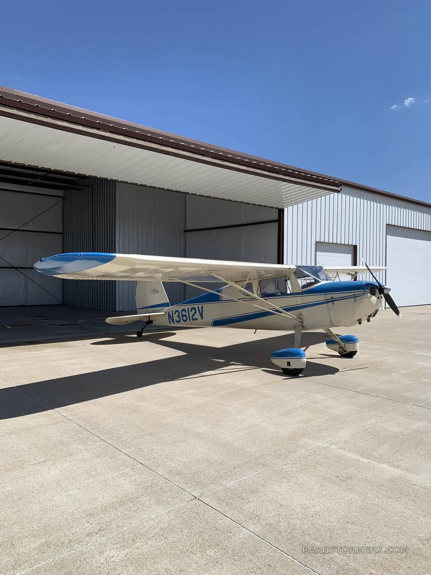 A white and blue Cessna 170B aircraft with tail number N3612V parked on a concrete pad outside a hangar under a bright blue sky.