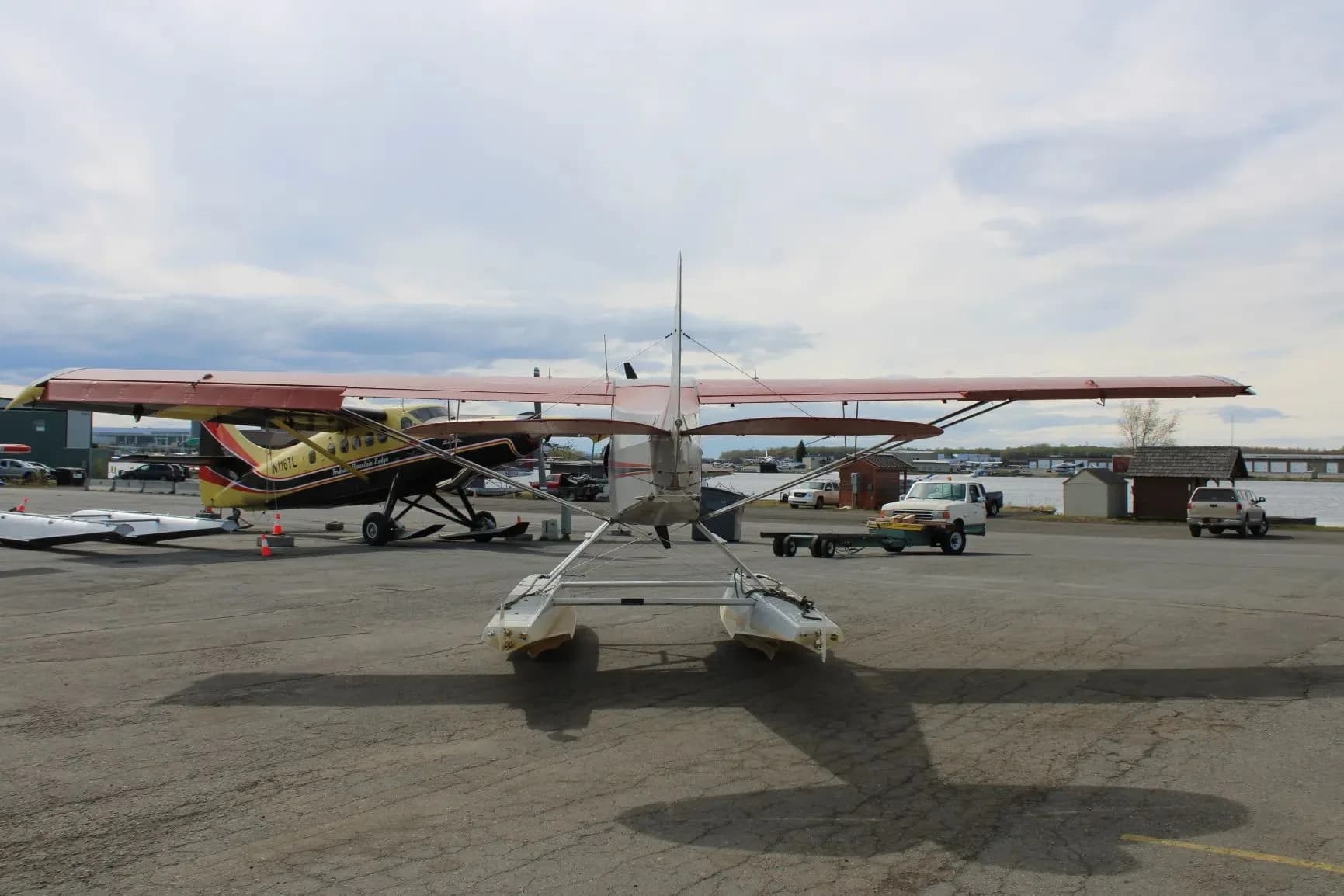 A rear view of the red and grey floatplane, showing its tail and floats, parked on an apron with other aircraft.