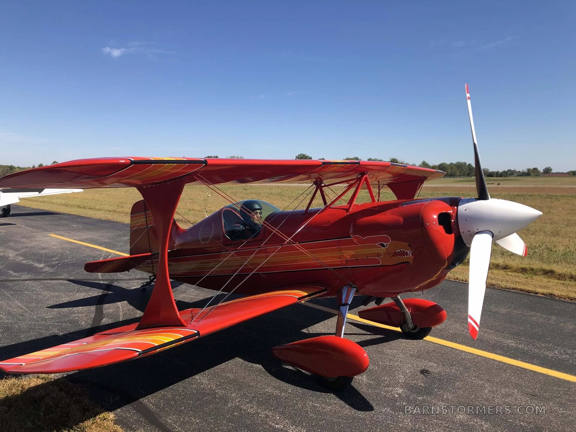A red biplane with orange and yellow stripes and a white propeller is parked on an airfield tarmac under a clear blue sky.