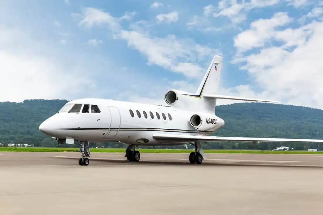 A Falcon jet sits on the tarmac with mountains in the background.