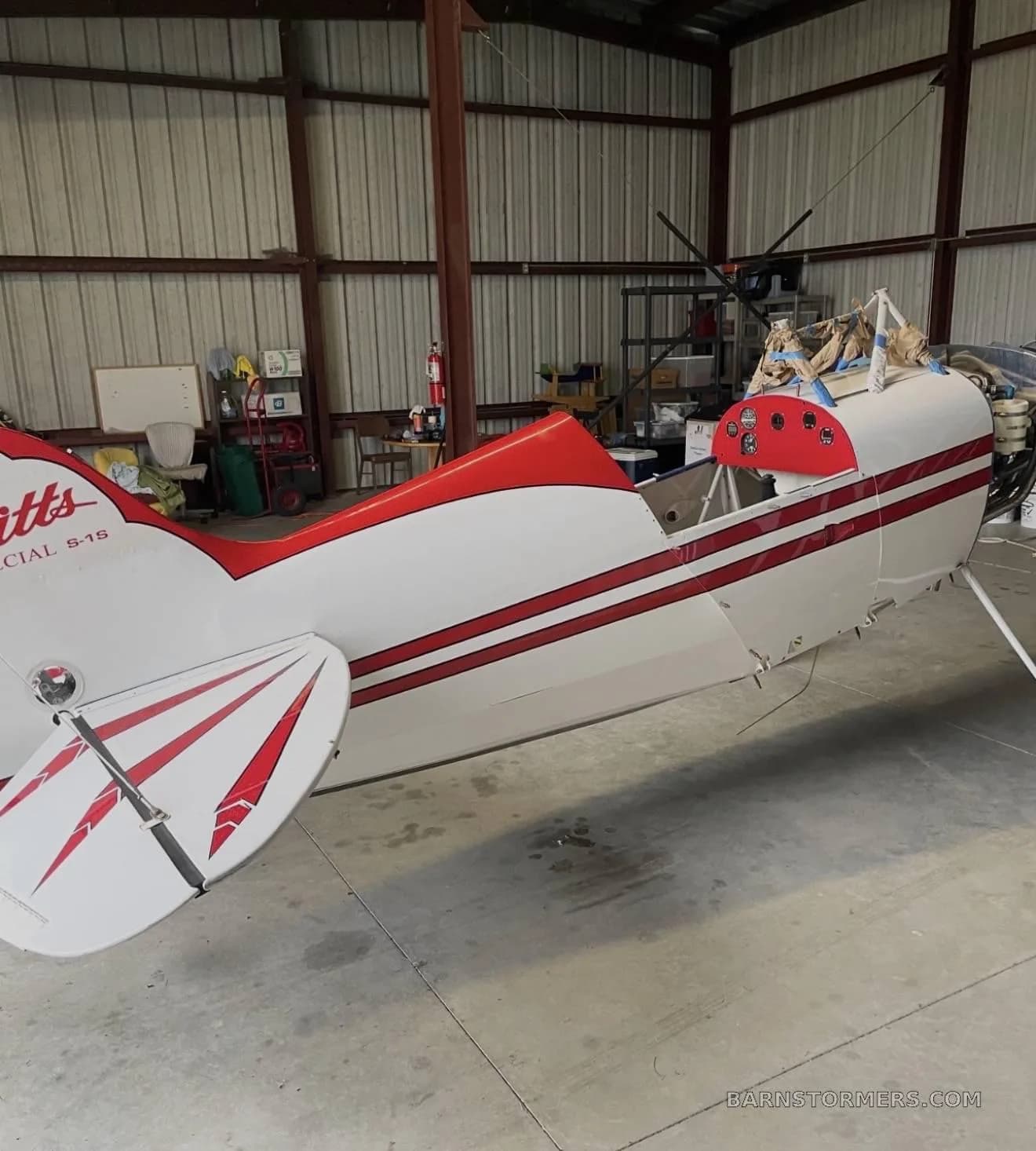 A close-up side view of the white and red Pitts Special fuselage and tail in a hangar.