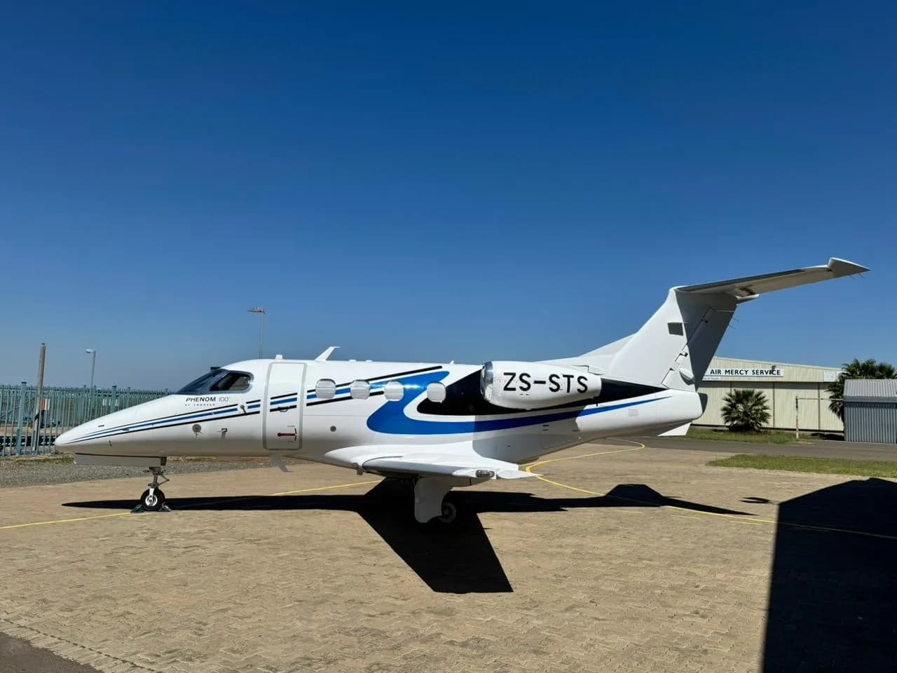 A white Embraer Phenom 100 with blue and black stripes (tail number ZS-STS) parked on an tarmac.