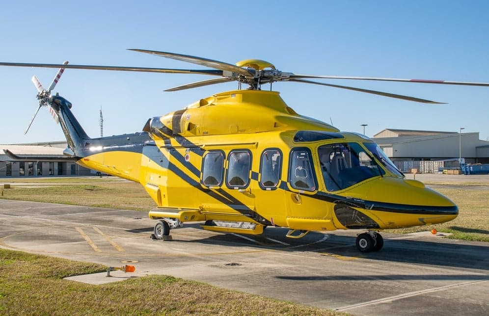 A bright yellow helicopter with black stripes is parked on an airfield with industrial buildings in the background.