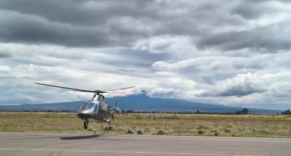 A grey AgustaWestland AW109 GrandNew helicopter is seen hovering just above a runway with a large mountain in the distance under a dramatic cloudy sky.
