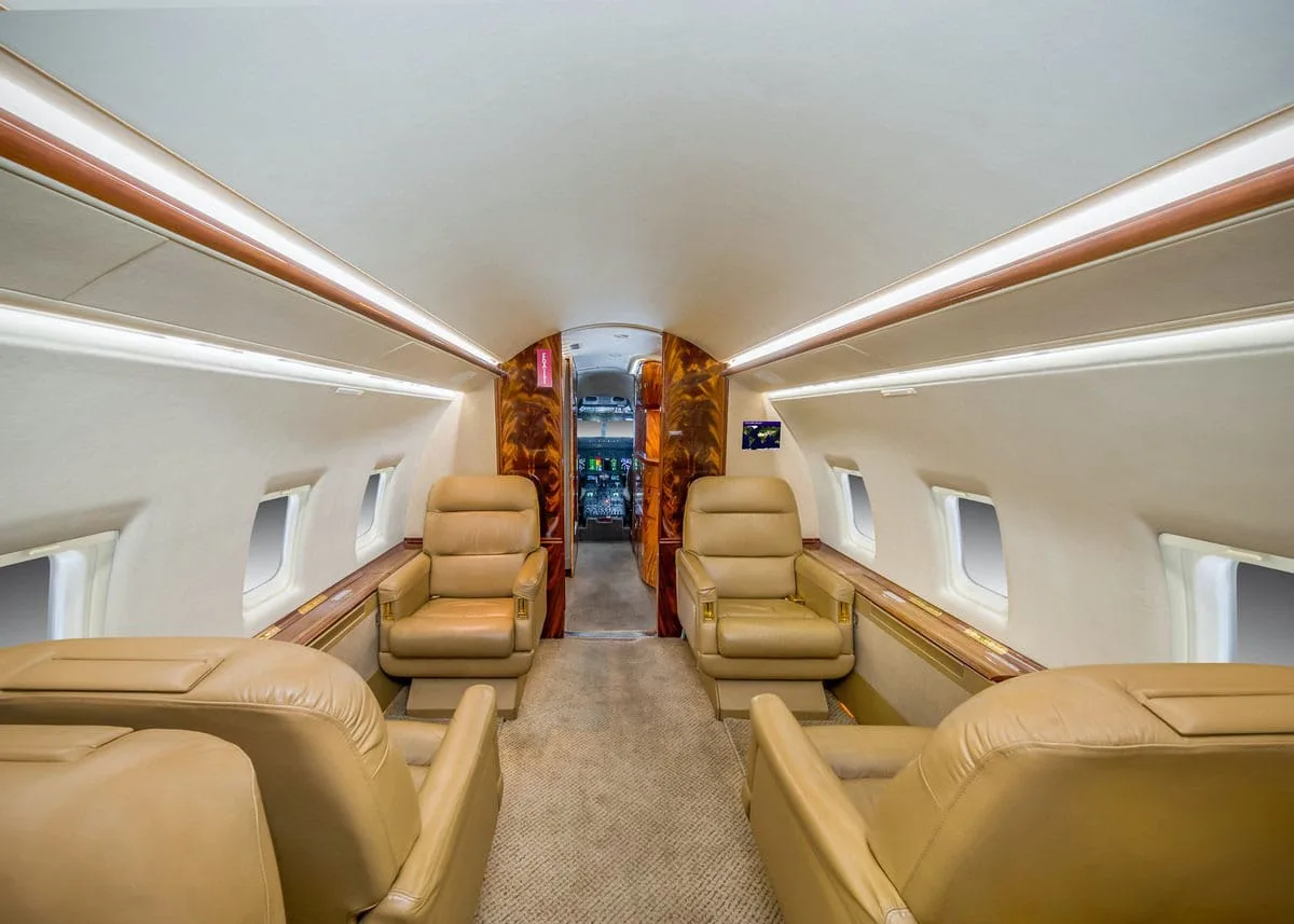 The spacious interior cabin of a business jet, featuring rows of tan leather seats facing each other and polished wood accents.