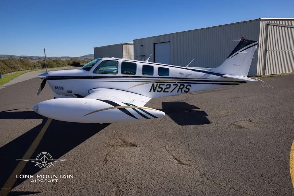 A white Beechcraft Baron N527RS with black and gold stripes parked on an asphalt next to hangars.