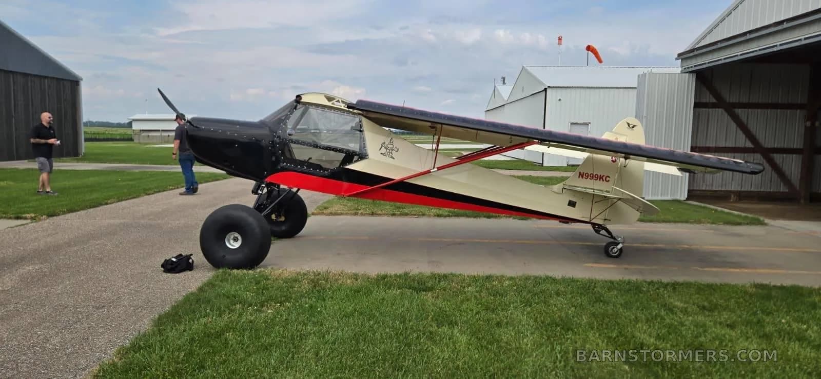 A black, red, and cream bush plane with large tires is parked on an asphalt ramp with hangars in the background and two men standing nearby.