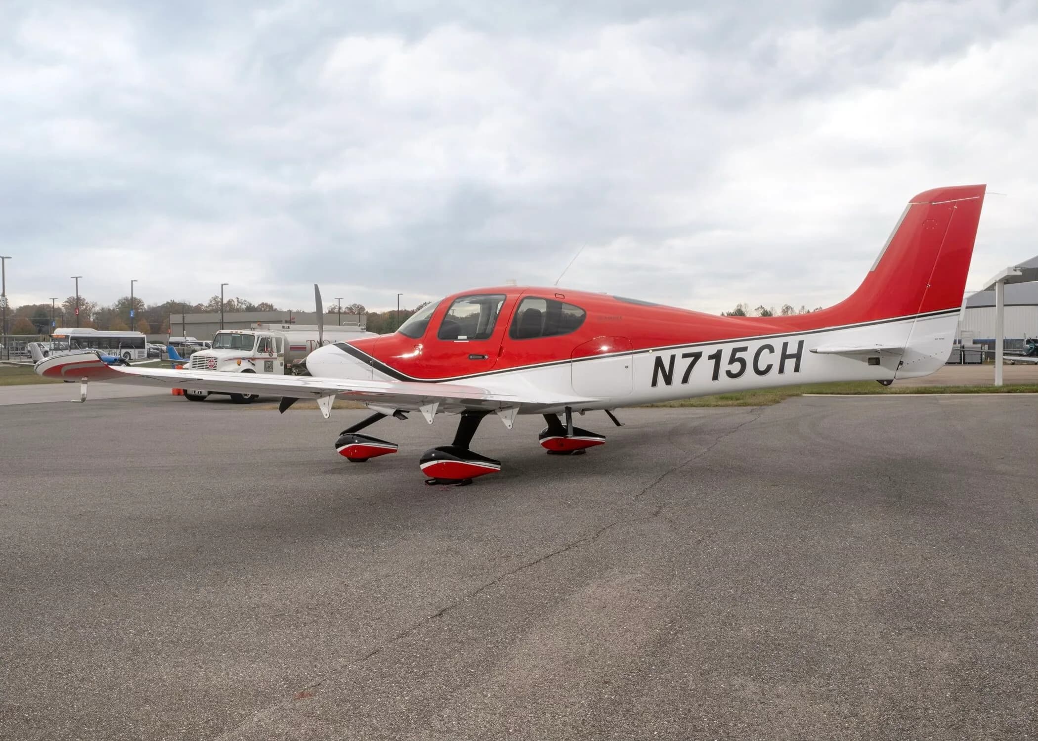 A red and white Cirrus SR22 aircraft with registration N715CH parked on an airfield with vehicles in the background.