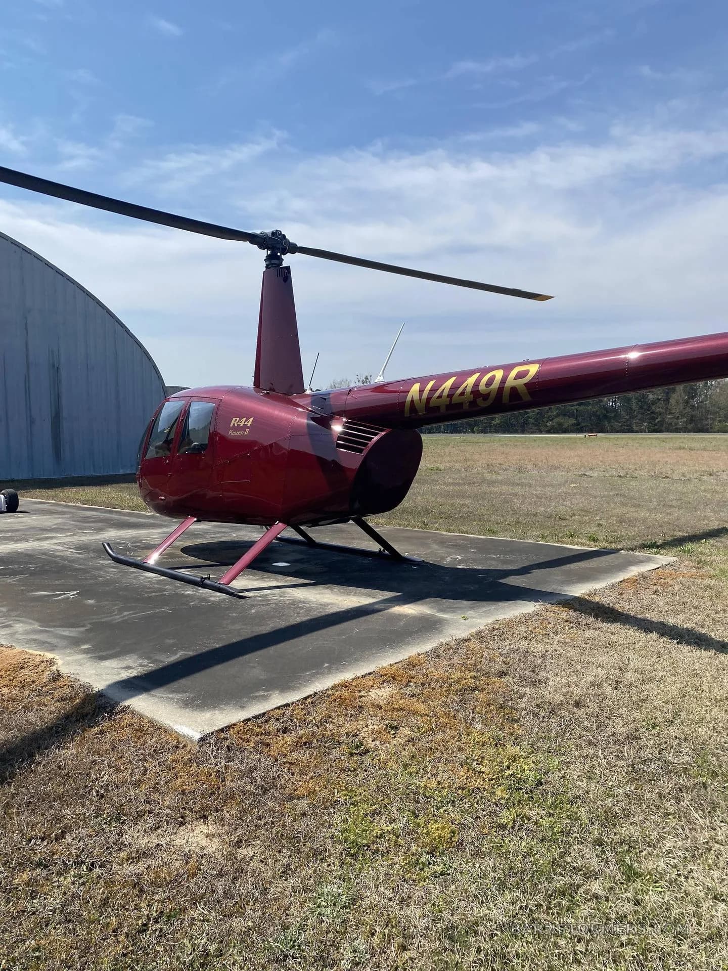 A dark red Robinson R44 Raven II helicopter with registration N449R, parked on a concrete pad next to a hangar.
