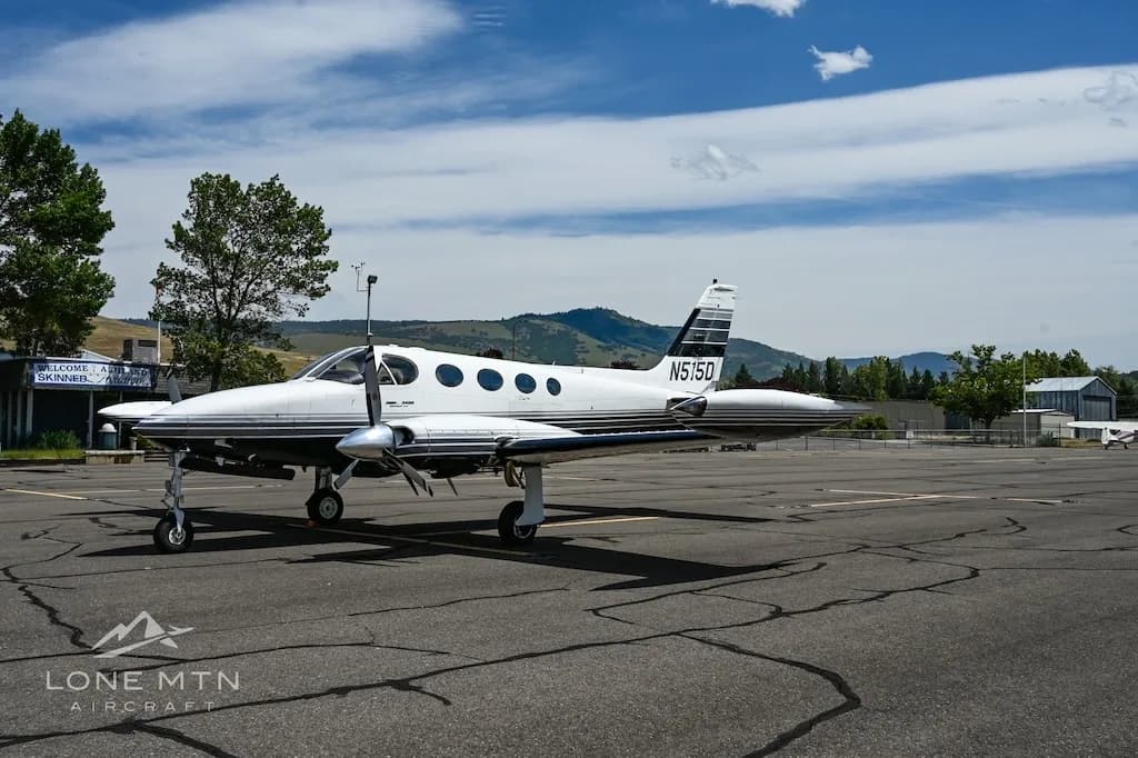 A side view of the white and black twin-engine aircraft, N515D, parked on an airfield next to trees and buildings.