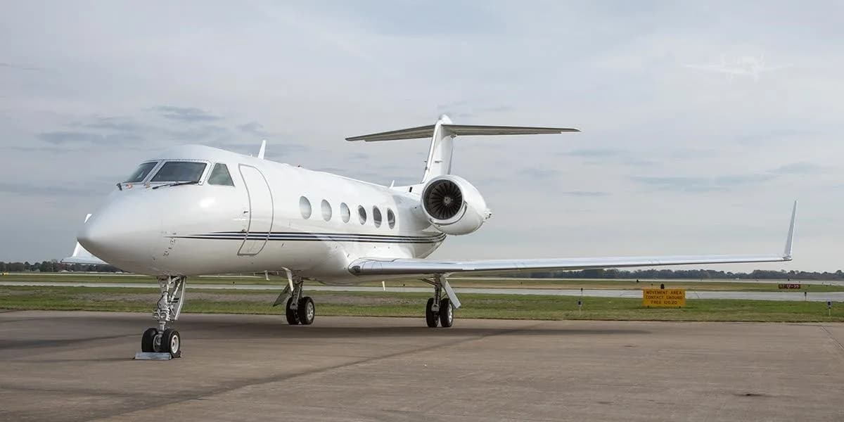 A white Gulfstream business jet with a thin dark stripe is parked on an airport tarmac under a cloudy sky.