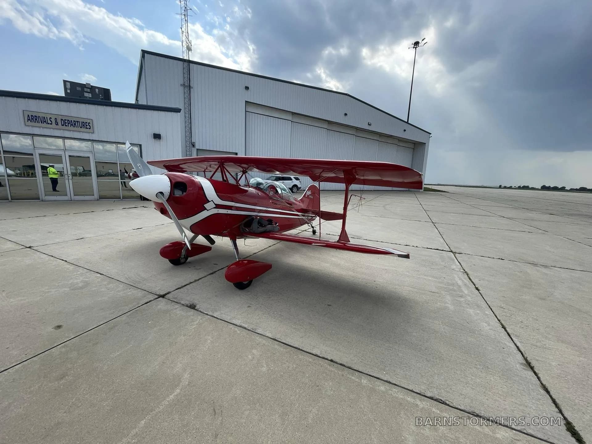 A red and white Pitts Special biplane is parked on an airfield tarmac next to a white hangar under a cloudy sky.