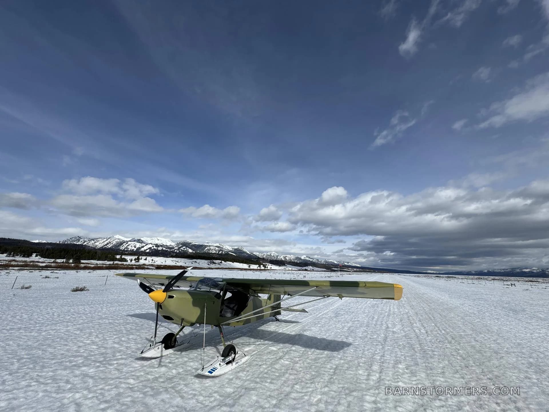 A camouflage-painted light aircraft with skis is parked on a snowy field with mountains in the background under a partly cloudy sky.